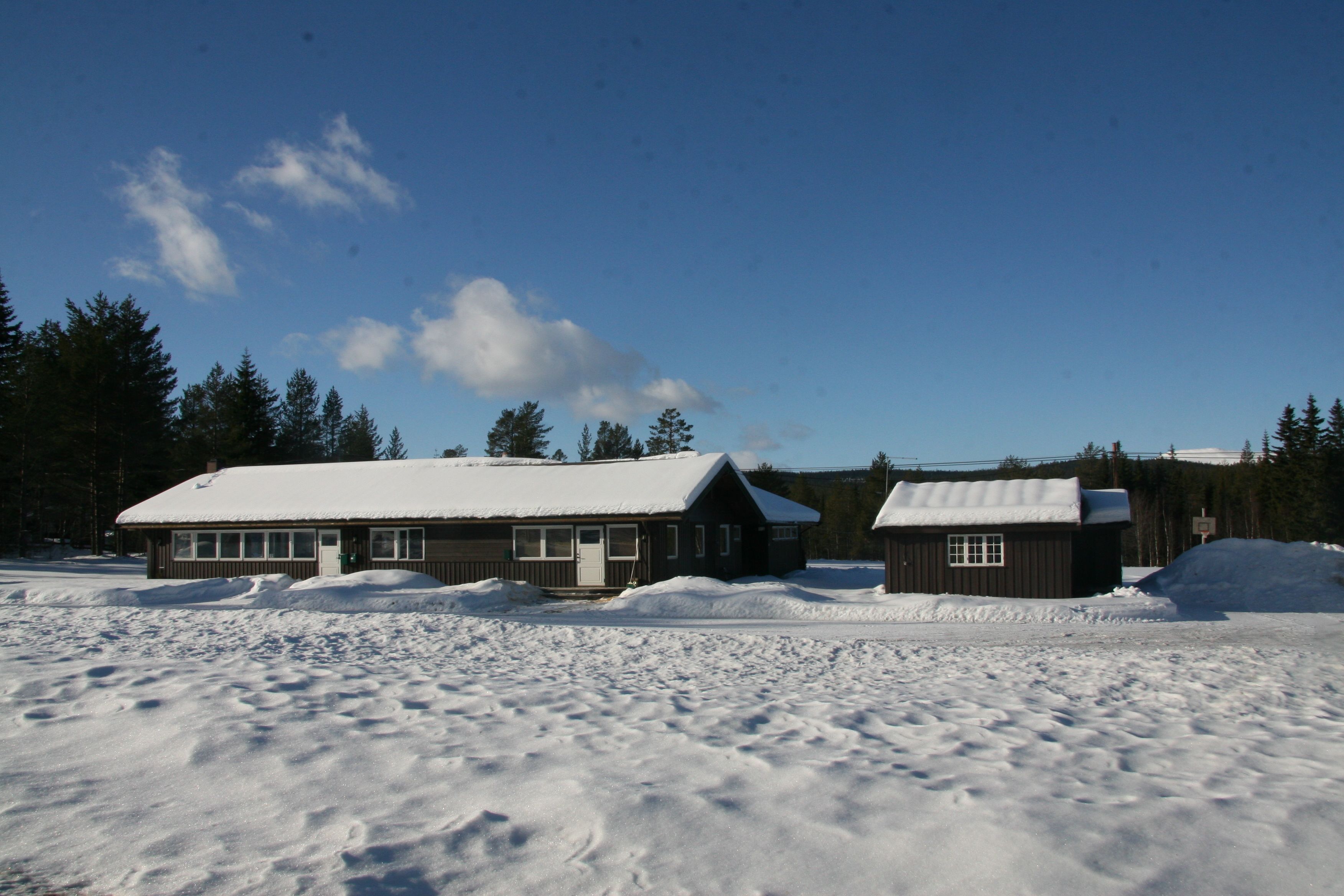Una cabana amb un teulat cobert de neu, envoltada per un paisatge nevós i una forest densa.