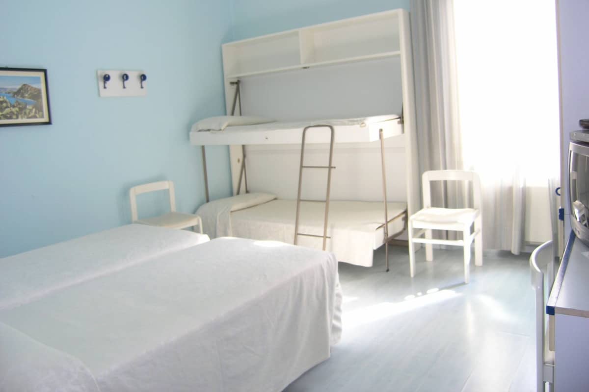 A room with a bunk bed, a single bed, two chairs, a desk, and a window with curtains.