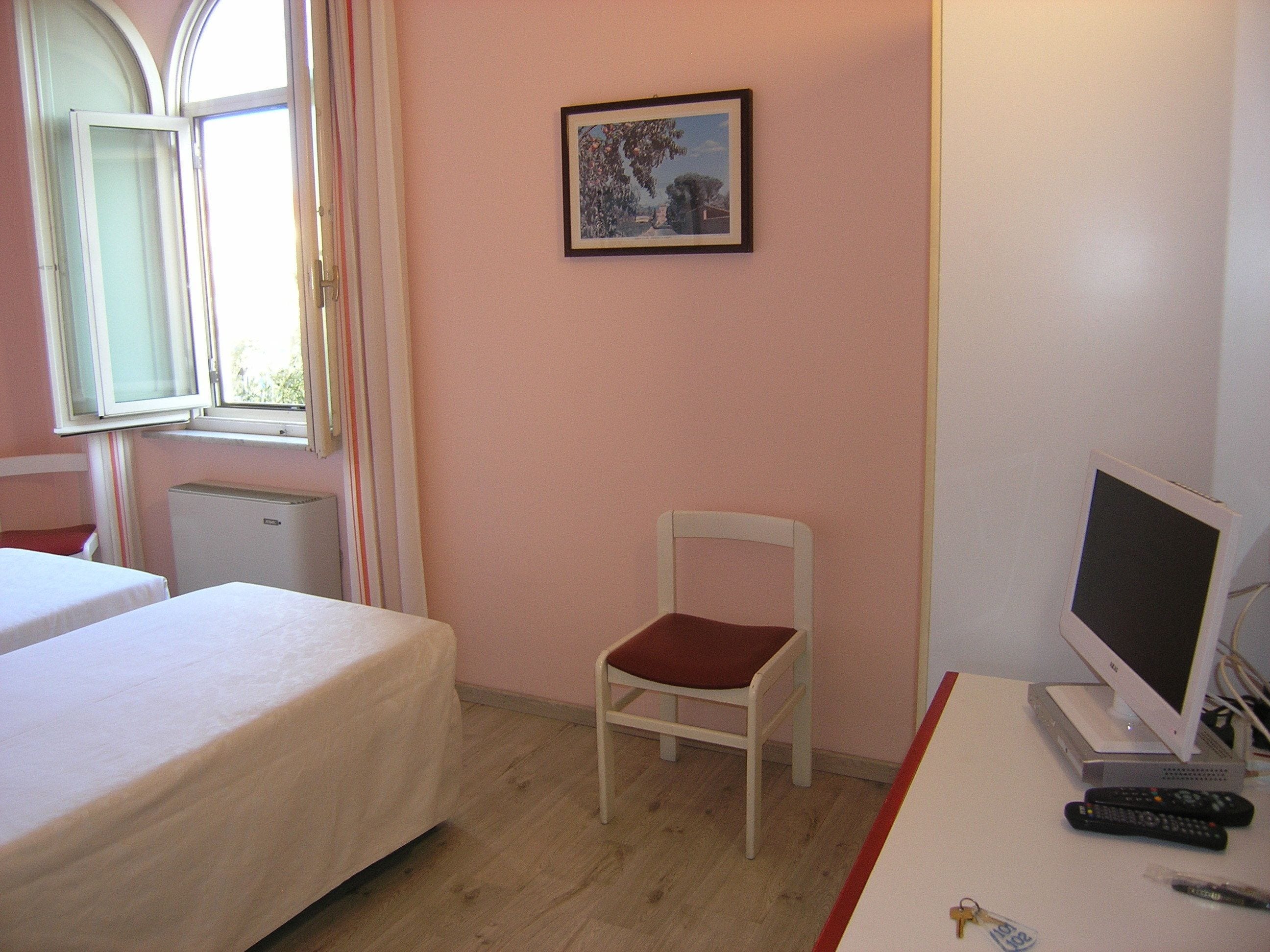 Habitación de hotel con dos camas, una silla, un escritorio con computadora y controles remotos, y una ventana con cortinas.