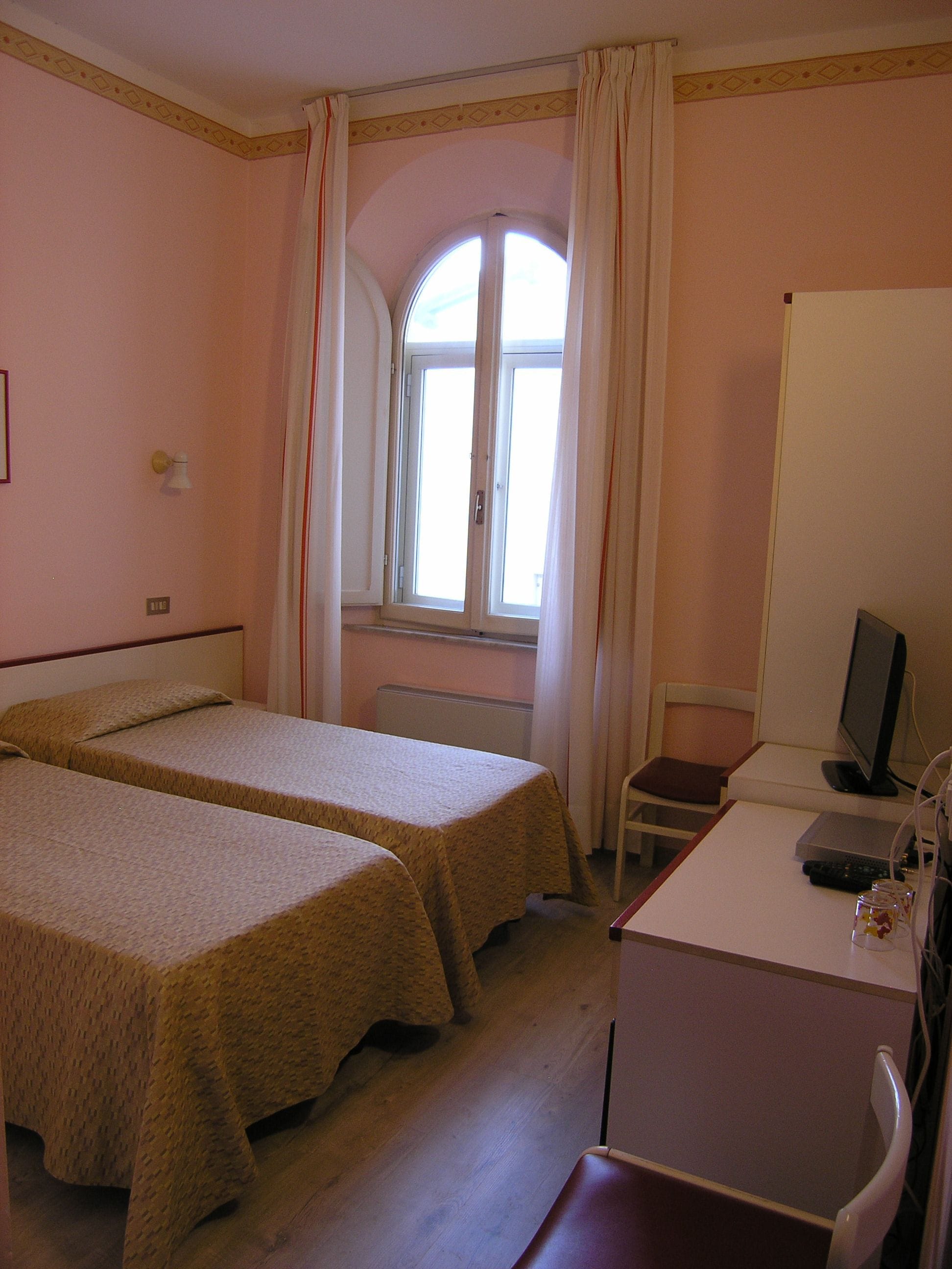 Ein Zimmer mit zwei Einzelbetten, einem Schreibtisch mit Computer, einem Stuhl und einem Fenster mit Vorhängen.
