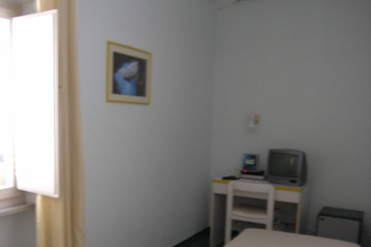 A small, compact room with a desk, a chair, a bed, a telephone, and a television.