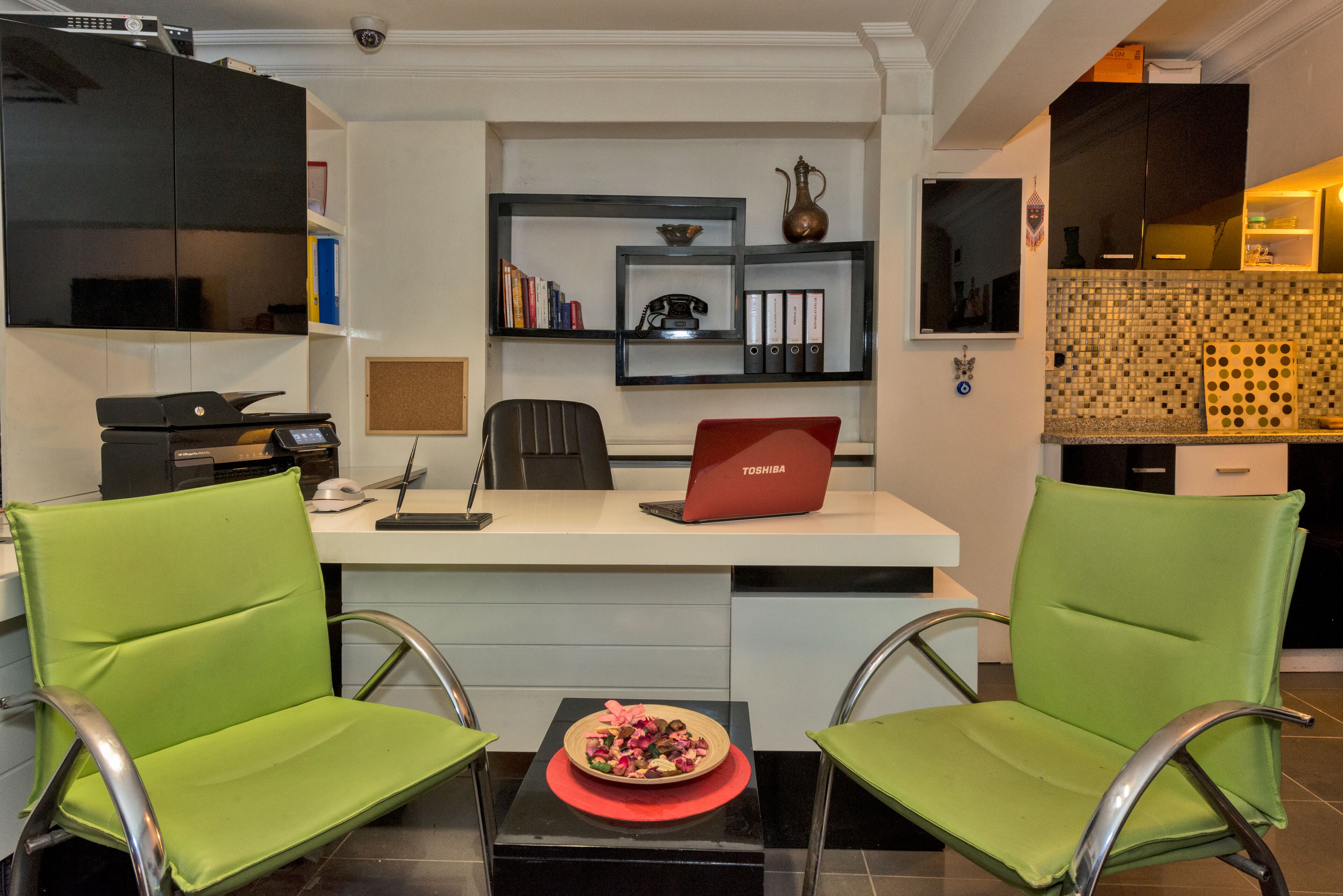 A modern kitchen with a refrigerator, microwave, and a red Toshiba laptop on a white countertop. There are two green chairs around a small table with a decorative bowl.