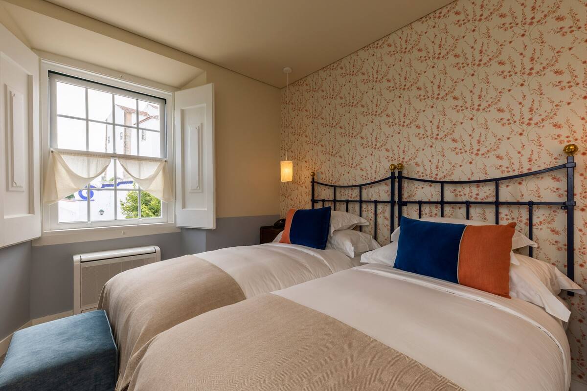 A bedroom with two beds, a window with curtains, a radiator, and a wall with floral wallpaper.