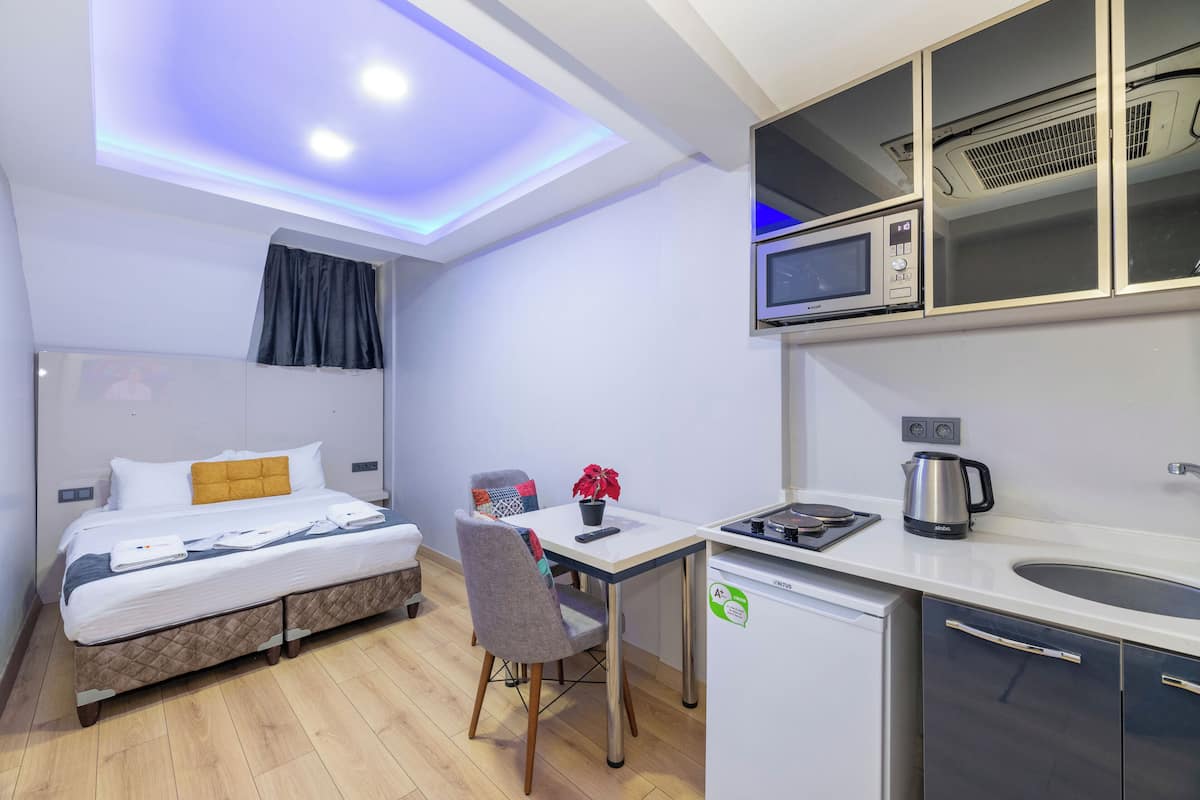 A compact hotel room with a bed, a small desk, a kitchenette, and a ceiling light.