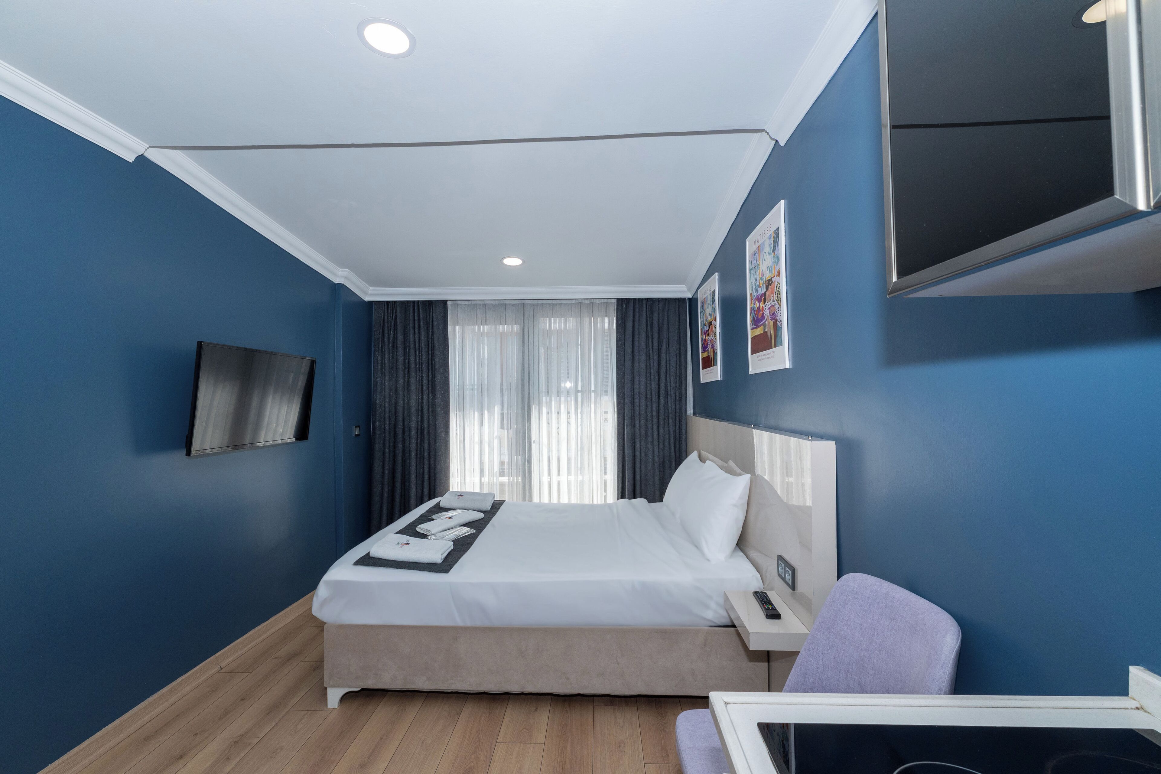 A modern hotel room with a large bed, a desk, a chair, a TV mounted on the wall, and a window with curtains.