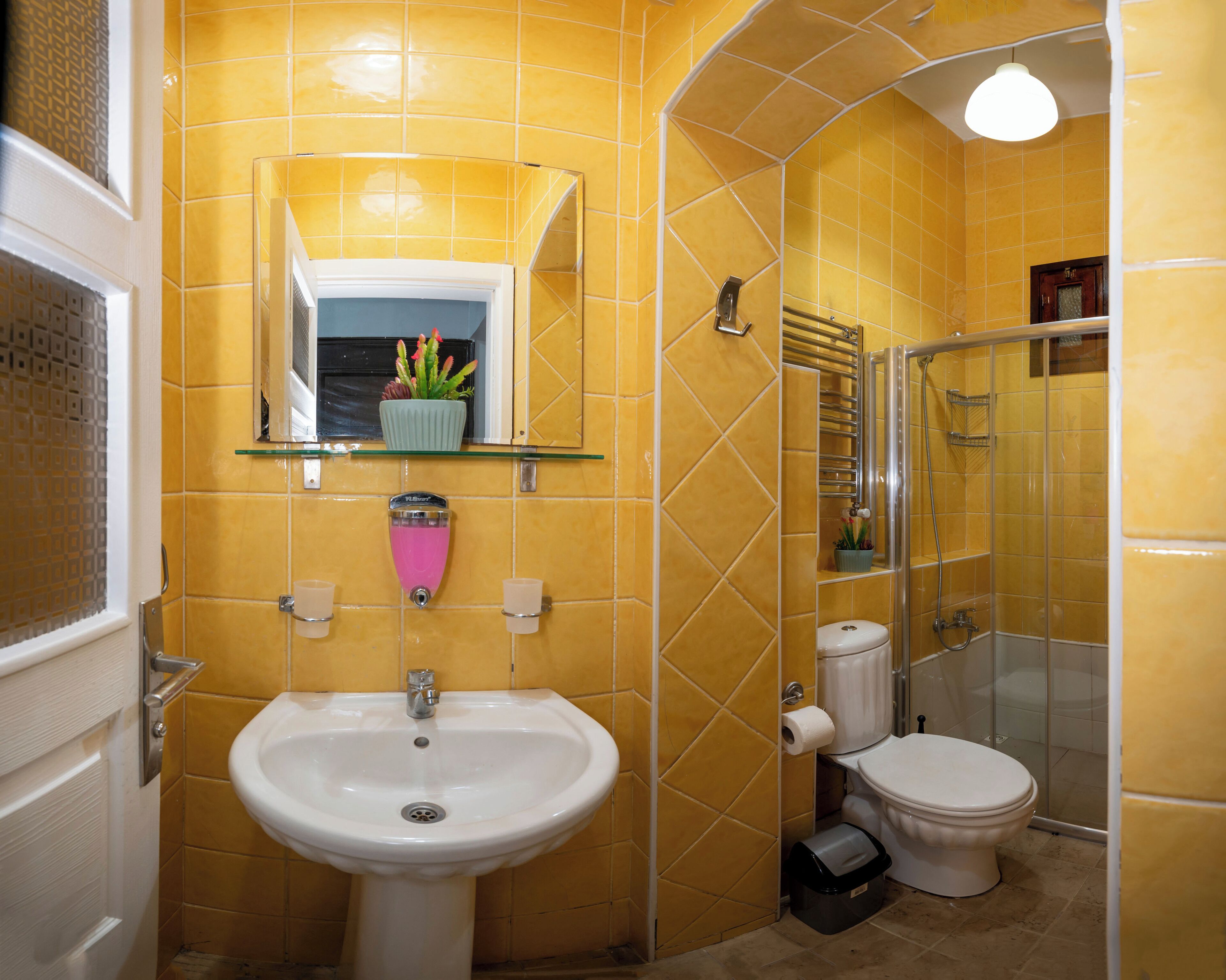 A bathroom with yellow tiles, a white sink, a toilet, a glass shower enclosure, and a mirror with a shelf above it.