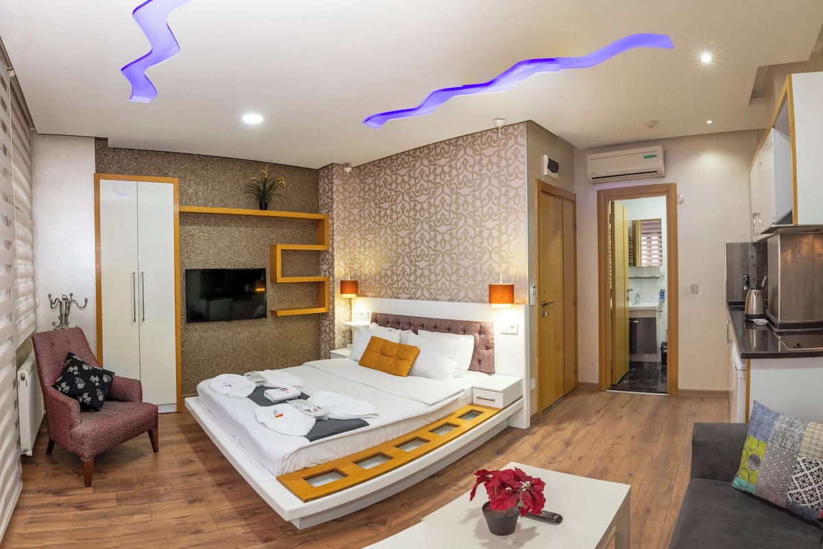 A modern hotel room with a bed, a sofa, a TV, and a kitchenette.