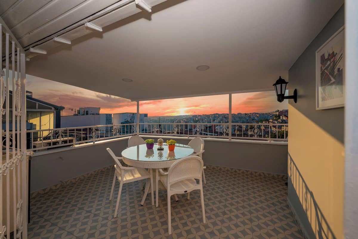 A round table with chairs and potted plants on a balcony with patterned flooring and a railing.