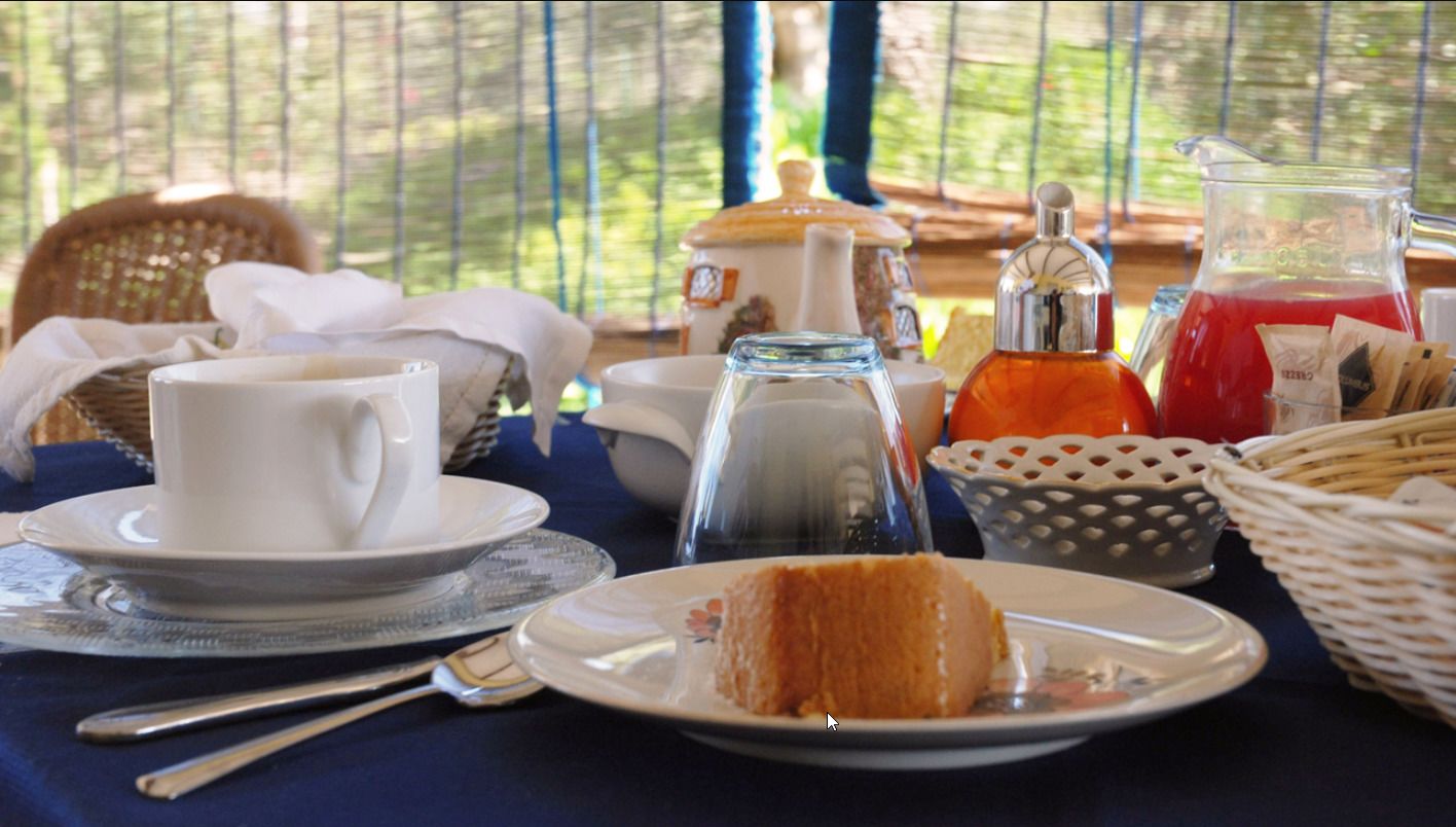 A table set with a white cup, saucer, and plate with a slice of cake, a glass pitcher, and a basket of tea bags.