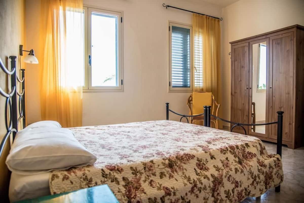 A bedroom with a bed, a chair, a wardrobe, and two windows with curtains.