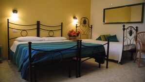 Ein Zimmer mit zwei Betten, jeweils mit Metallgestell und einer Matratze. An beiden Seiten der Betten befinden sich Wandlampen, und über dem rechten Bett ist ein Spiegel angebracht. Ein Korbstuhl steht in der Nähe des rechten Bettes.