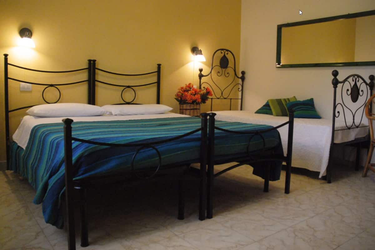 A room with two beds, each with a metal frame and a single mattress. There are wall-mounted lamps on either side of the beds, and a mirror above the right bed. A wicker chair is positioned near the right bed.