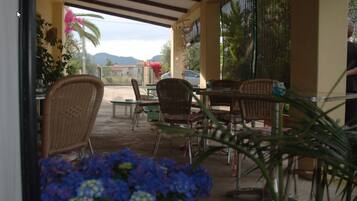 Un espace extérieur aménagé en terrasse, avec des meubles en osier, une table et des chaises. On y trouve des plantes en pot et, au loin, une vue sur les montagnes.