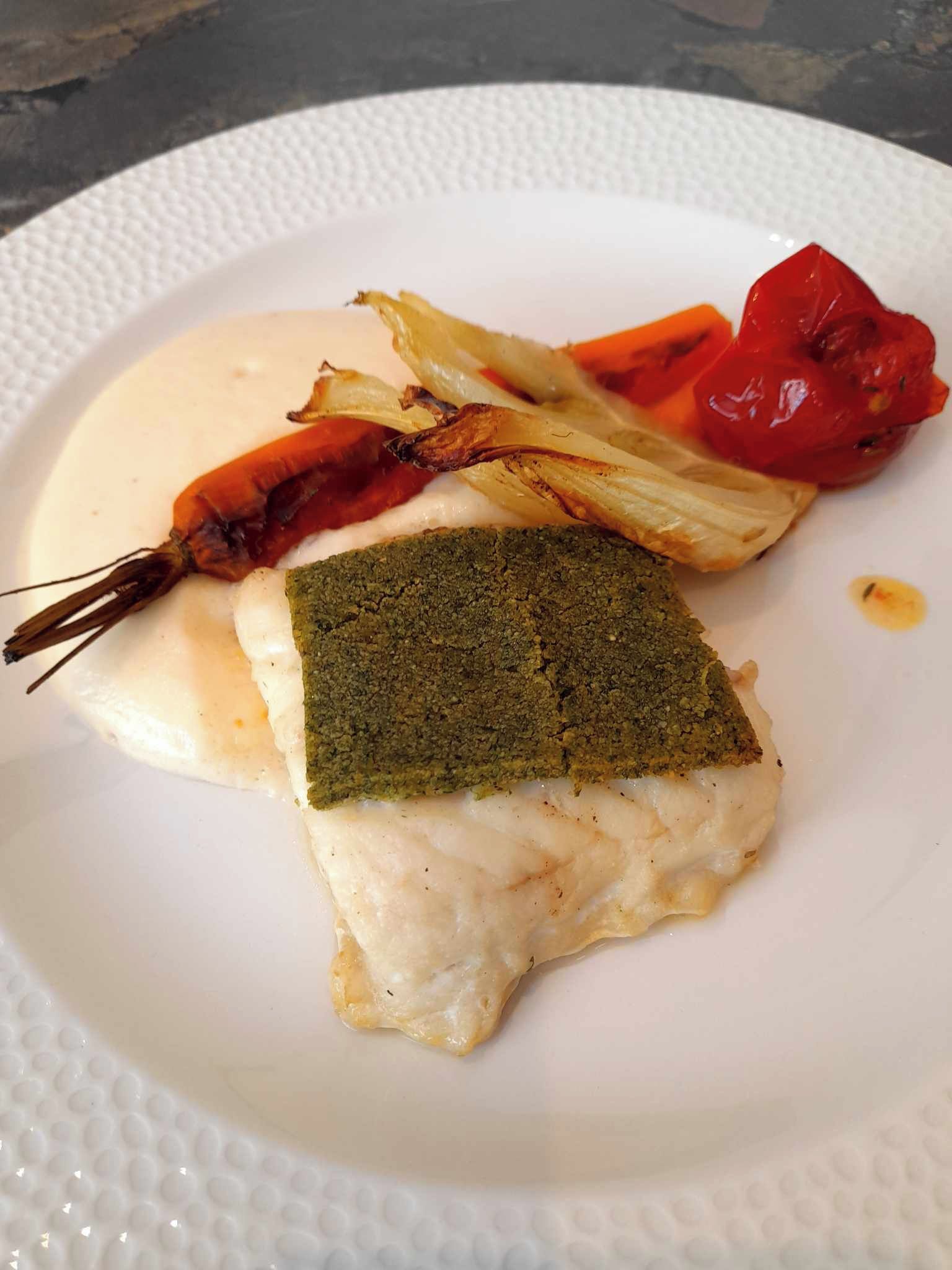 Une assiette blanche sur laquelle on trouve un morceau de poisson garni d’herbes vertes, une crevette grillée et une tomate rouge.