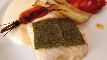 Une assiette blanche sur laquelle on trouve un morceau de poisson garni d’herbes vertes, une crevette grillée et une tomate rouge.