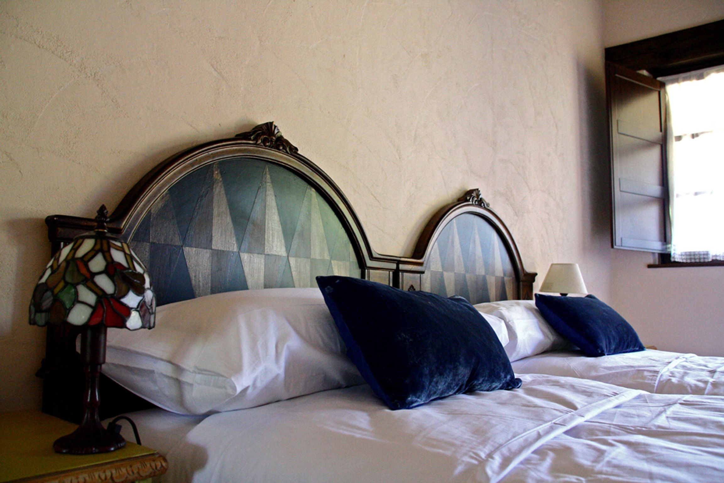 A bed with white linens and dark blue pillows, a wooden headboard, a stained glass lamp, and a bedside table.