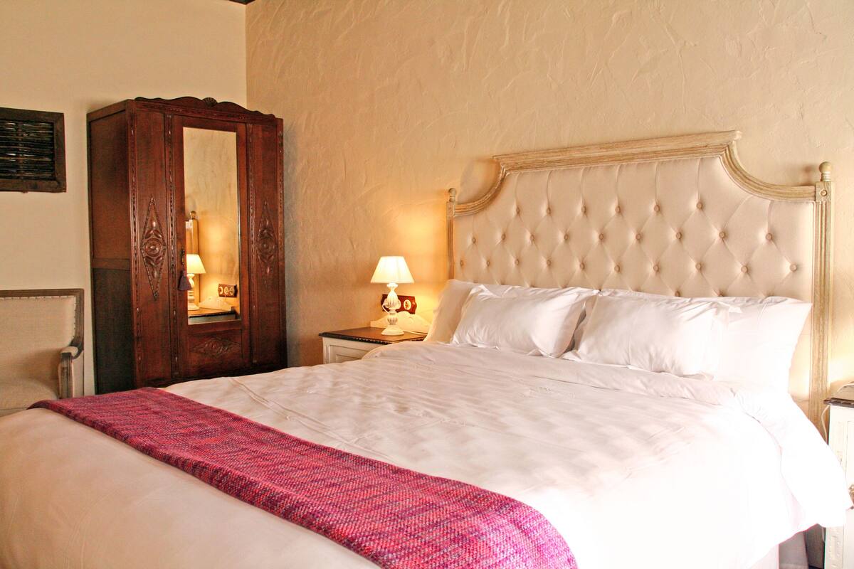 Una cama bien hecha con edredón blanco y manta rosa, una mesita de noche de madera con lámpara, y un espejo grande en la pared.