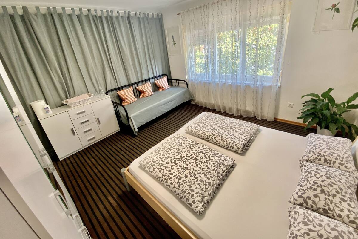 Une chambre à coucher comprenant un lit, une table de chevet, une commode, une fenêtre avec des rideaux et une plante en pot.