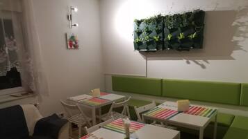 Een kamer met een groen gestoffeerde bank, witte tafels met kleurrijke tafelkleden met strepen, en een plantenplank aan de muur.