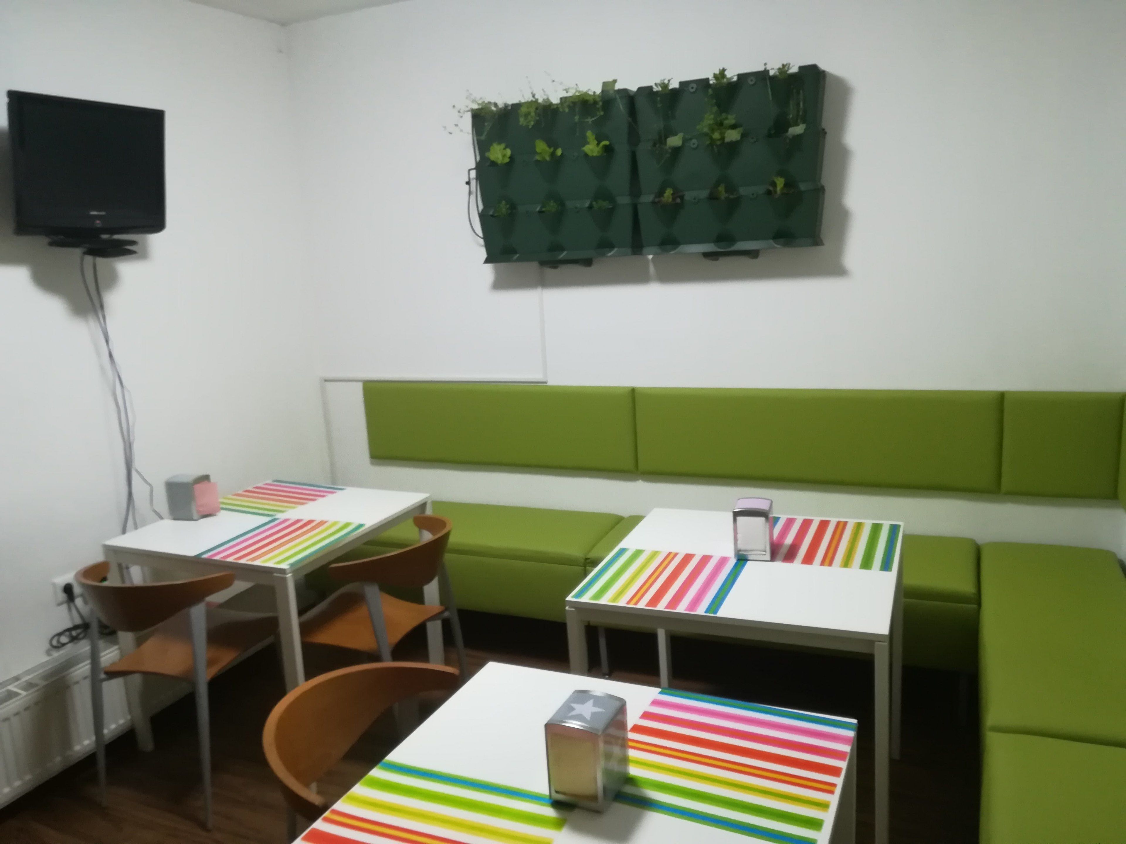 A room with a green padded bench, two tables with striped tablecloths, a television mounted on the wall, and a green plant wall hanging.