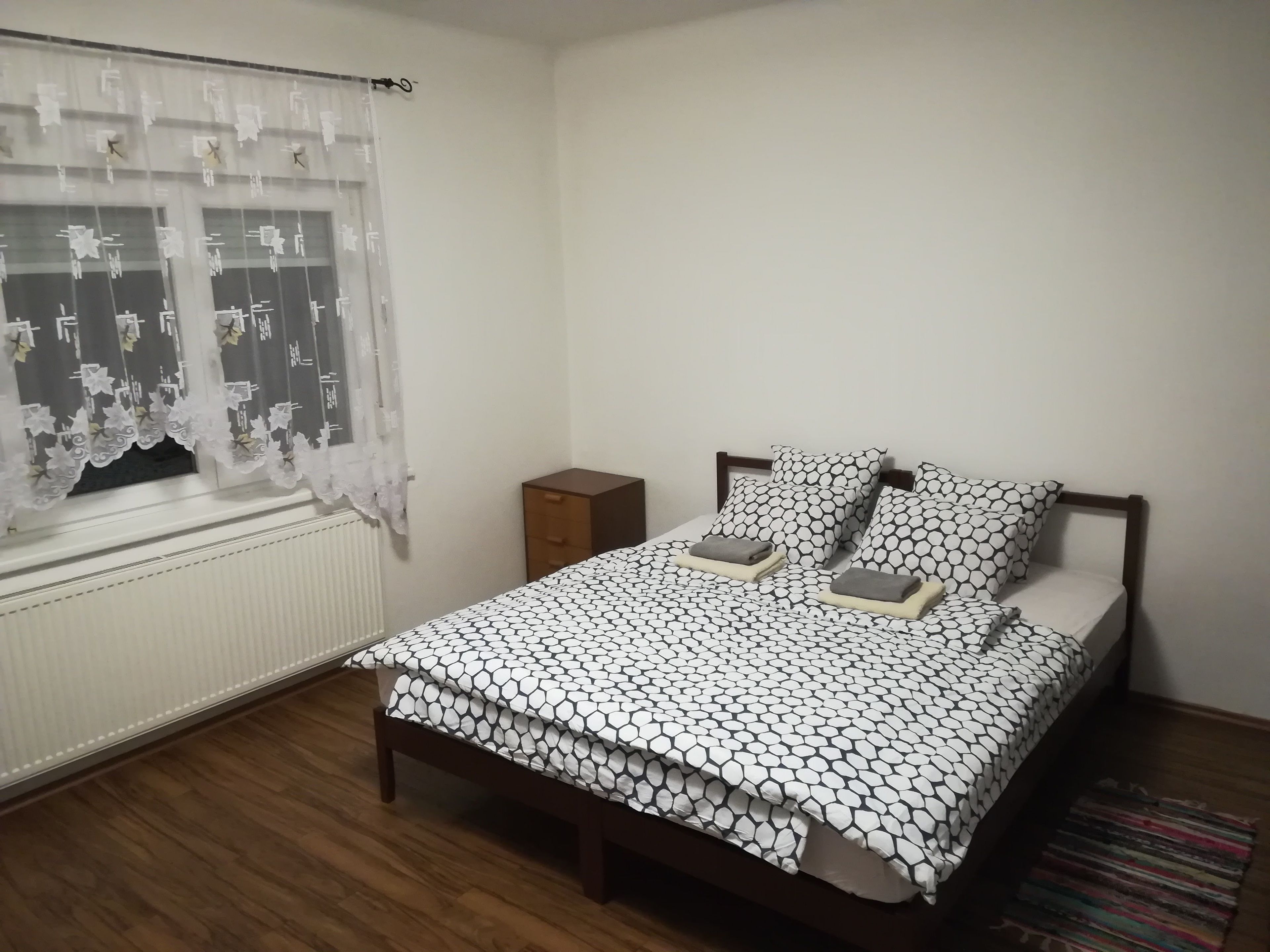 Un dormitorio con cama, mesita de noche de madera, ventana con cortina translúcida y radiador.