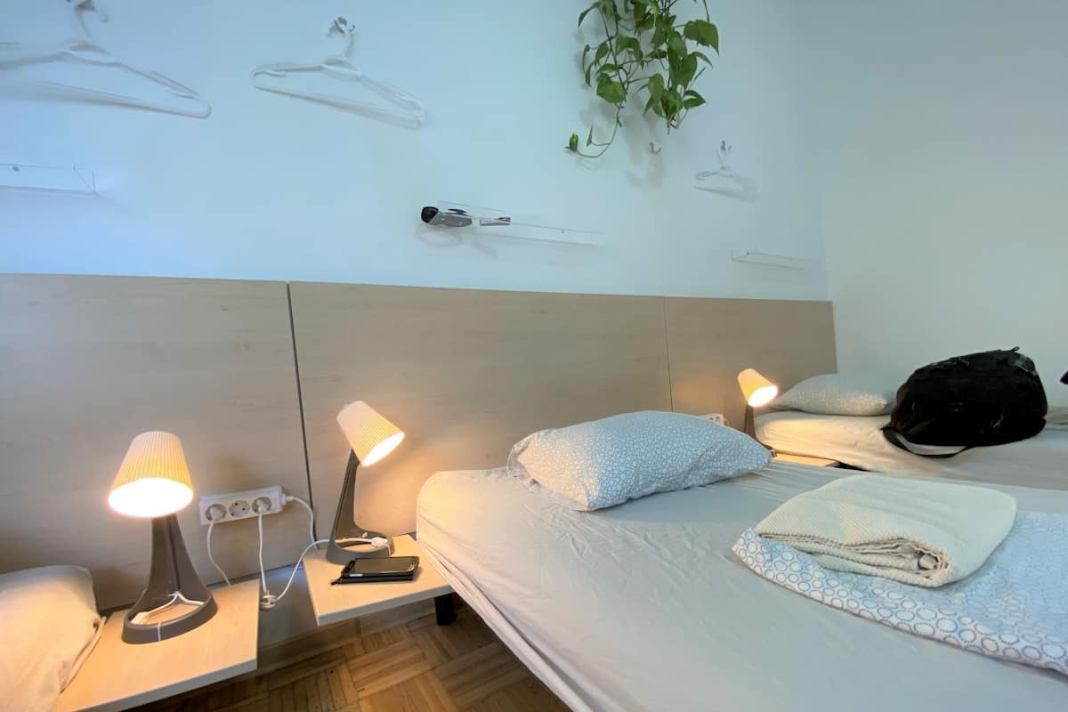 Une chambre d’hôtel avec un lit, des lampes de chevet, une table de chevet avec un téléphone, une plante fixée au mur et un sac à dos posé sur le lit.
