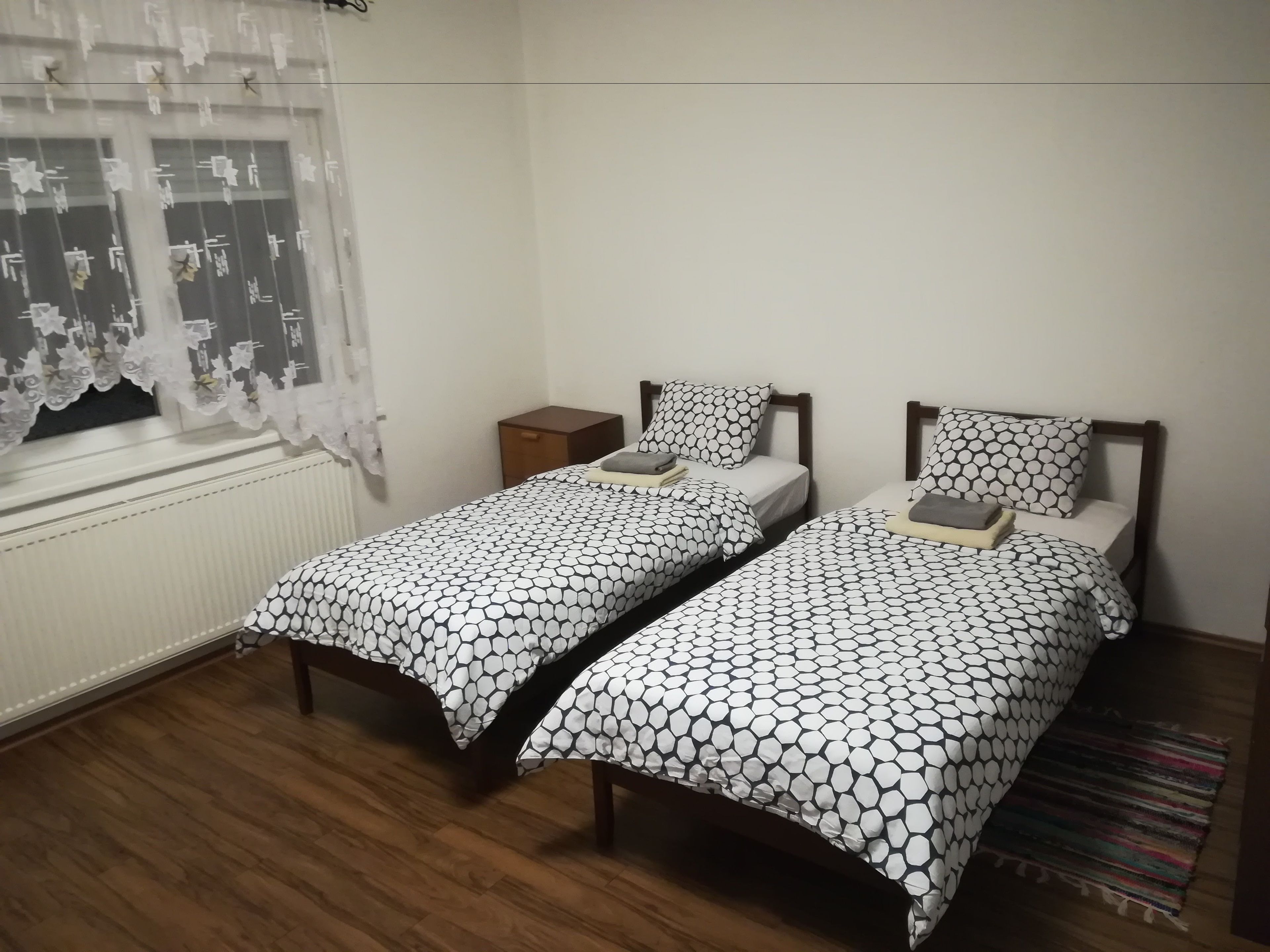 A room with two single beds, a small wooden nightstand, a window with lace curtains, and a wooden floor.