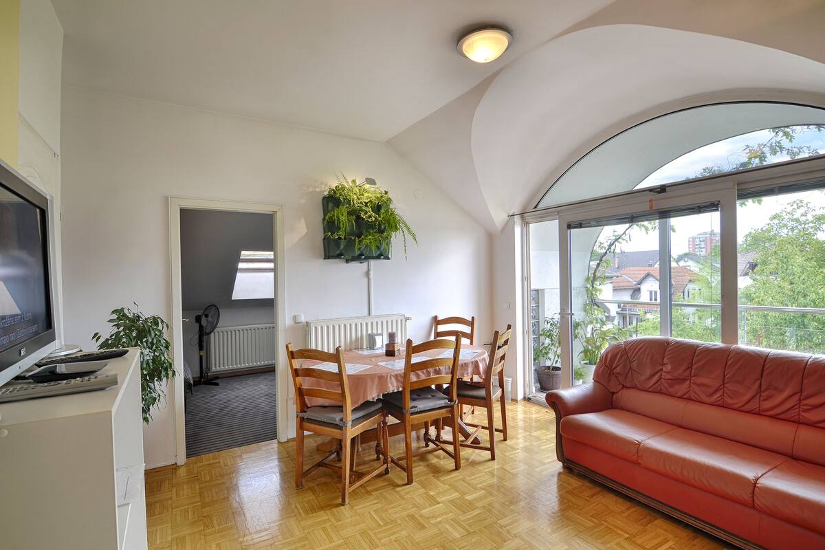 Un coin repas avec une table et des chaises, un canapé rouge et une grande fenêtre donnant sur des immeubles et des arbres.