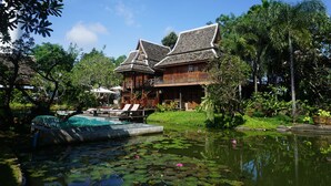 A traditional wooden house with a swimming pool surrounded by lush greenery and a pond with lily pads. - Baansuwanburi (San Sai)