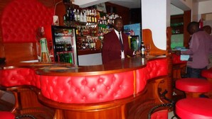 Una zona de bar con un mostrador y taburetes acolchados de color rojo, una estantería bien surtida con licores, y una persona de pie detrás del bar.