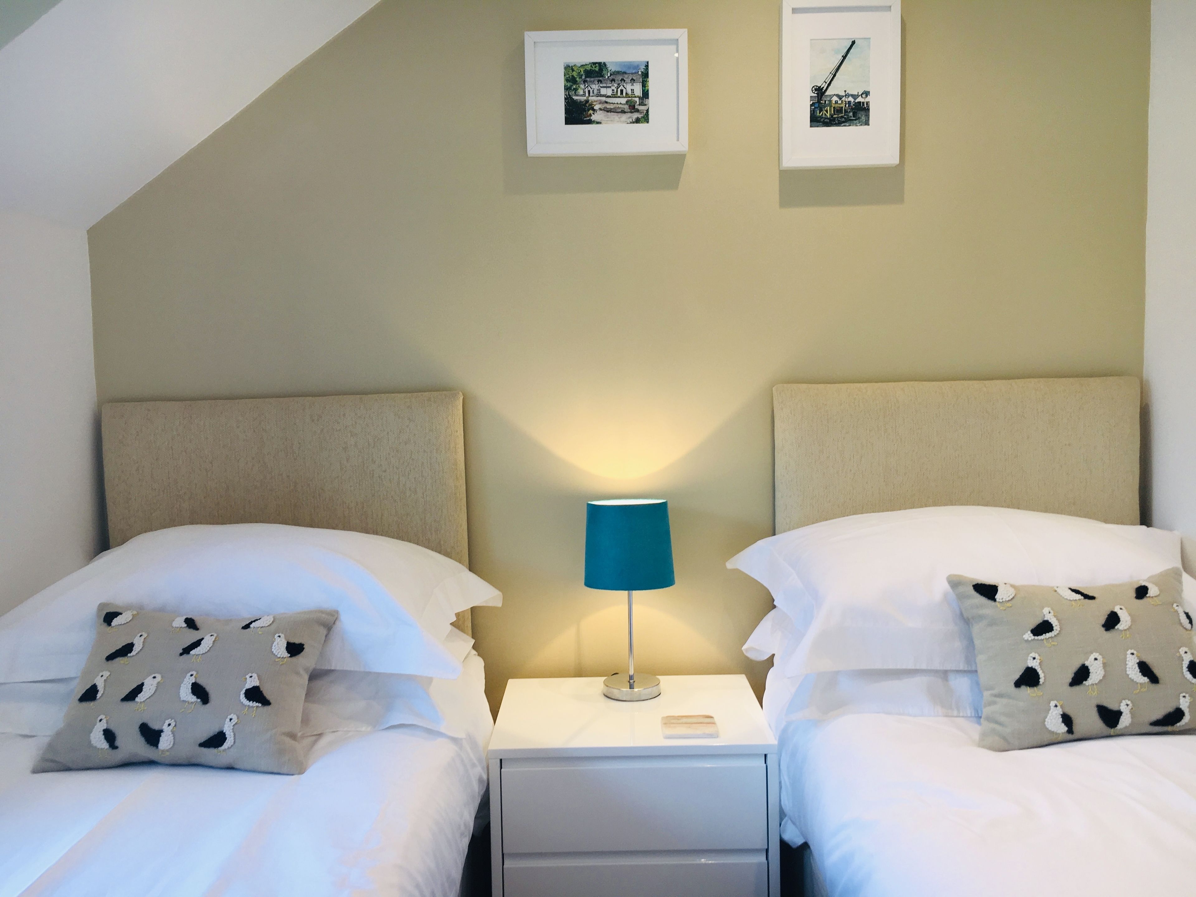 Habitación de hotel con dos camas, cada una con un cojín estampado con un diseño de pájaros. Hay una mesita de noche con una lámpara entre las camas.