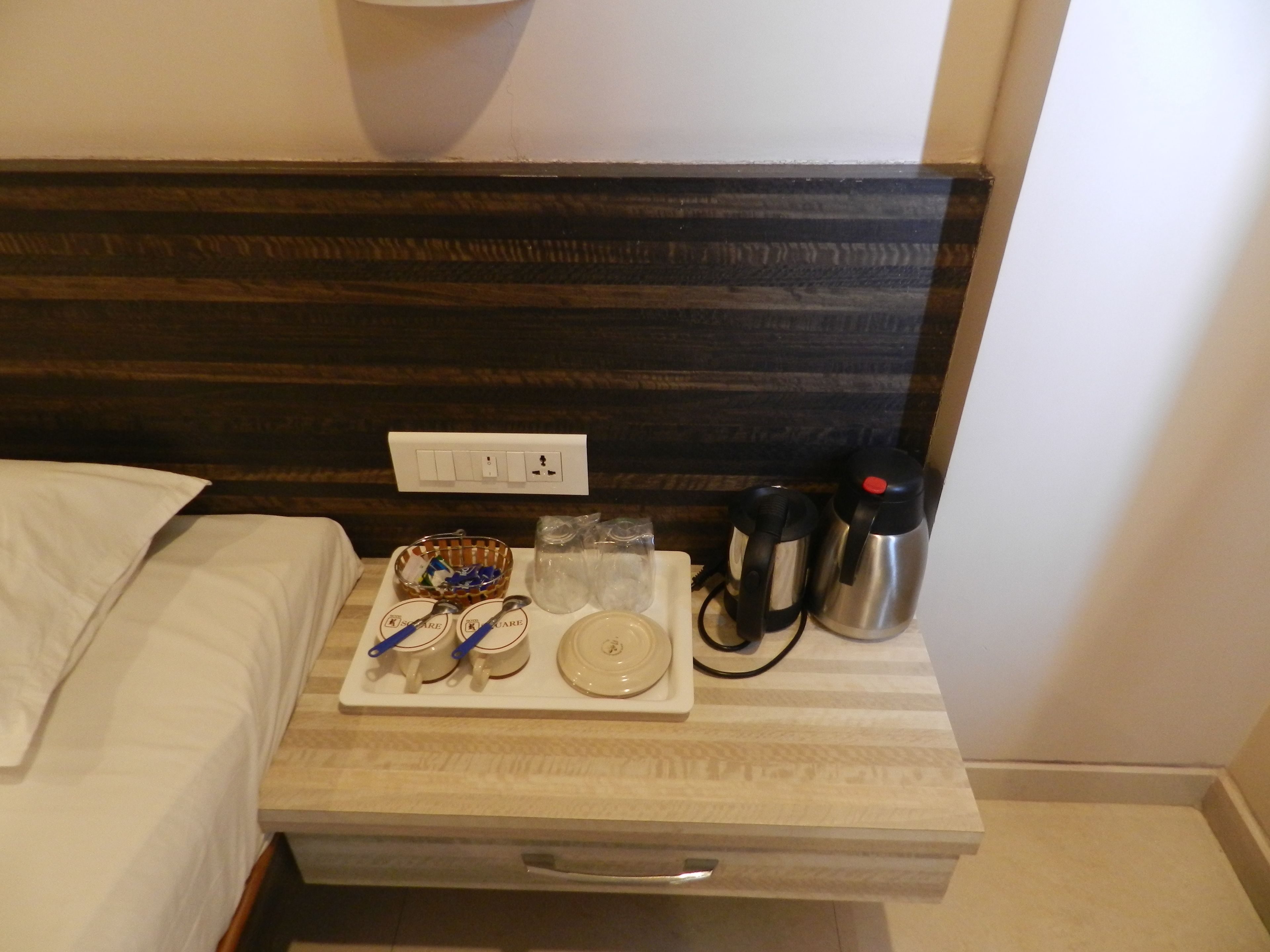 A hotel room with a bed, a bedside table with a tray of breakfast items, a kettle, and a power outlet.