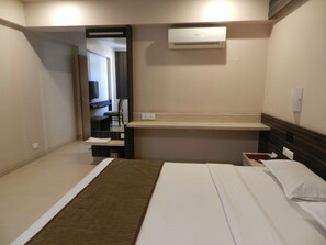 A hotel room with a bed, a bedside table, a small desk with a chair, a mirror, and an air conditioning unit.