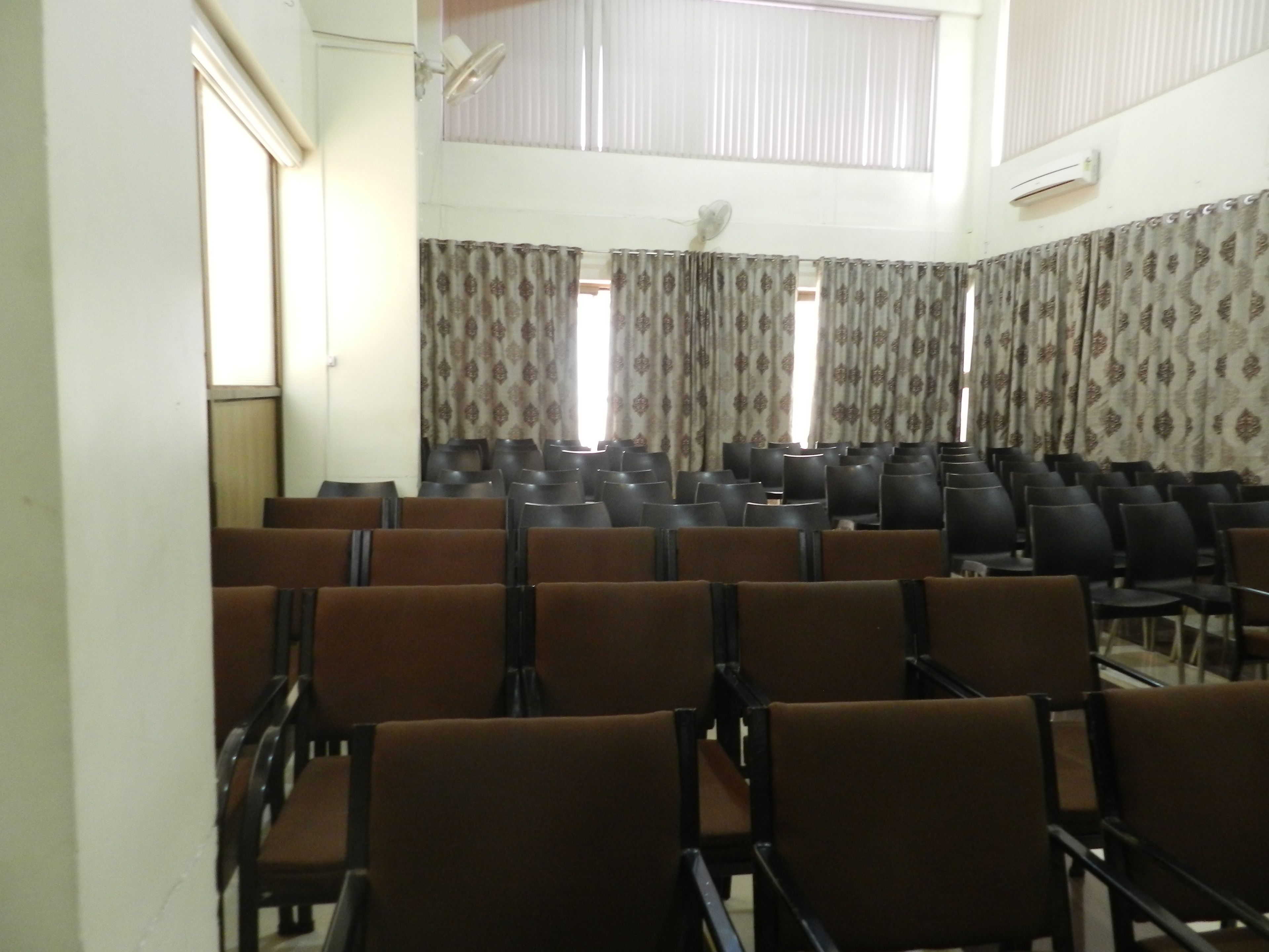 A conference room with rows of chairs, patterned curtains, and an air conditioning unit on the wall.