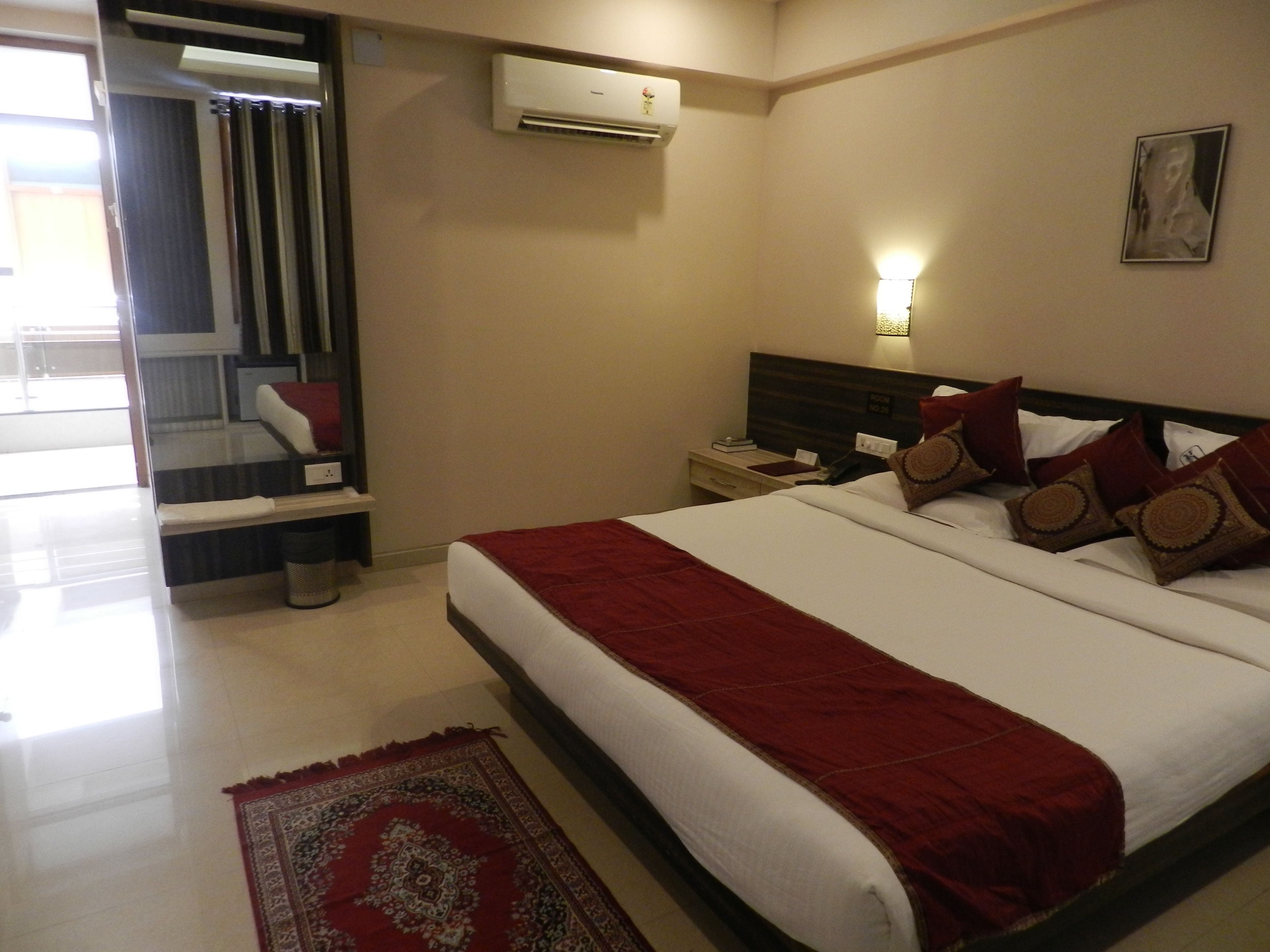 Habitación de hotel con una cama grande, una mesita de noche, un equipo de aire acondicionado, una lámpara de pared y una puerta que da a un balcón.