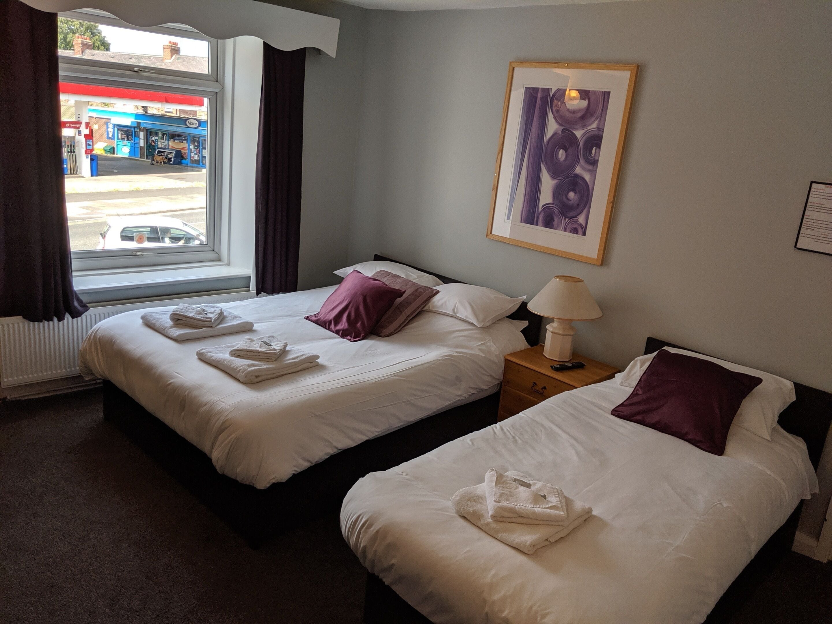 Habitación de hotel con dos camas, una ventana con cortinas, un radiador, un cuadro en la pared y una mesita de noche con lámpara.