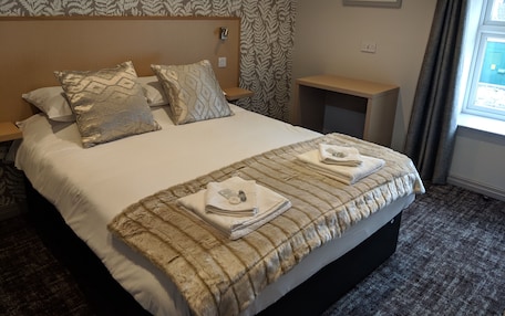 A neatly made bed with a striped blanket and pillows, a wooden headboard, a small bedside table, and a window with curtains.