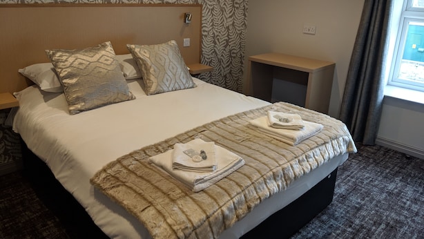 A neatly made bed with a striped blanket and pillows, a wooden headboard, a small bedside table, and a window with curtains.