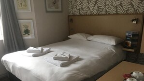 A neatly made bed with white linens and two folded towels, a wooden nightstand with a tray of white cups and a red packet, and a patterned wallpaper above the bed.