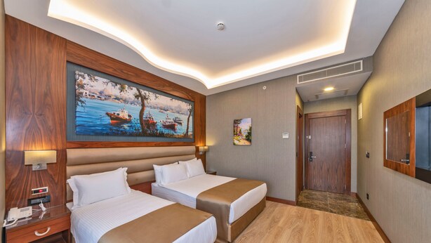 A hotel room with two beds, a painting of a boat on water, a wooden headboard, a desk with a lamp, and a door leading to another room. - Piya Sport Hotel (Istanbul)