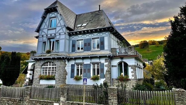 Une maison à deux étages, dotée d’un toit à forte pente, d’une façade en pierre et de nombreuses fenêtres équipées de volets.
