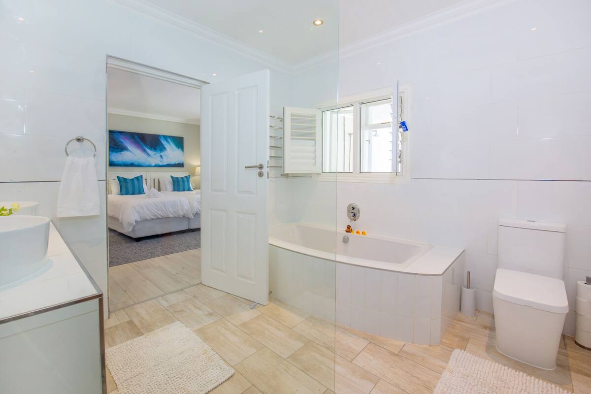 A modern bathroom with a bathtub, toilet, and sink. There is a glass shower enclosure and a bed visible through an open door.