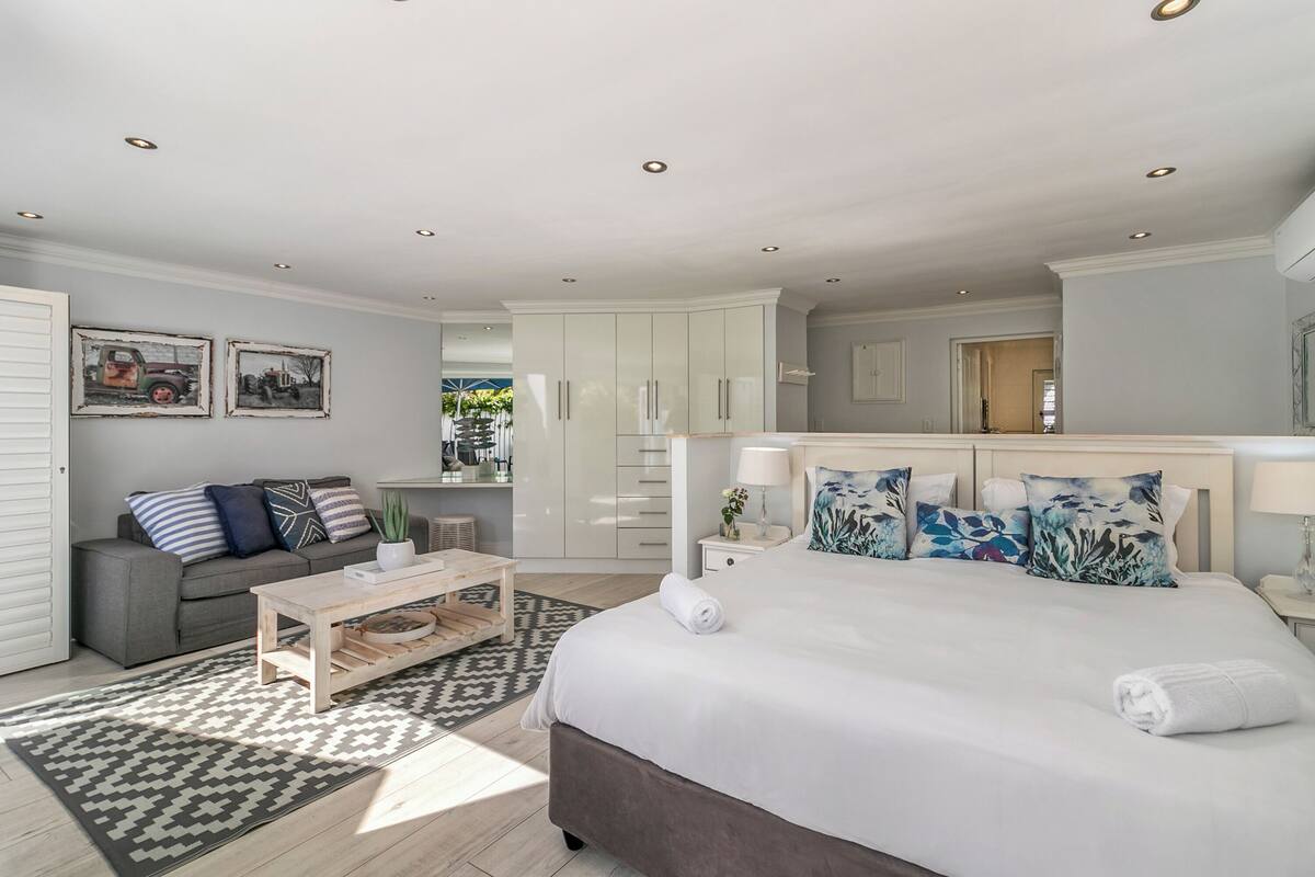 A spacious bedroom with a large bed, a grey sofa, a wooden coffee table, and a patterned rug.