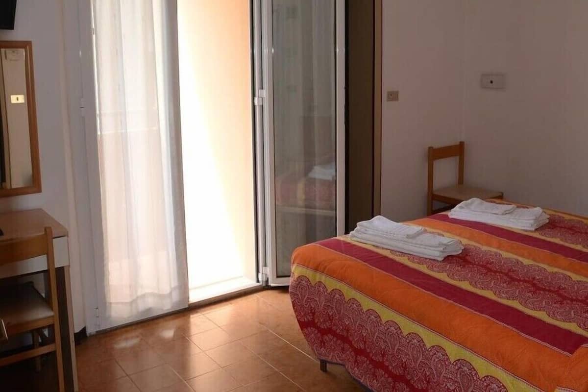 A bedroom with a bed, a chair, a desk, and a sliding glass door leading to a balcony.
