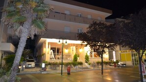 Hotel de vários andares com entrada bem iluminada, escadaria visível e área de estacionamento.