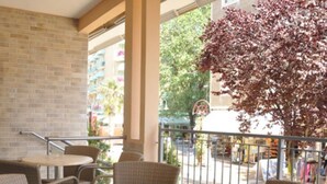 Uma área de estar coberta ao ar livre com mobiliário de vime e uma mesa de vidro, com vista para uma rua com edifícios e uma árvore com folhas vermelhas.