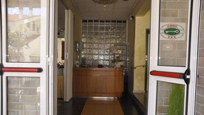 Uma área de recepção de hotel com um balcão de madeira, divisória de vidro e uma placa com a inscrição 'ALBERGO'.
