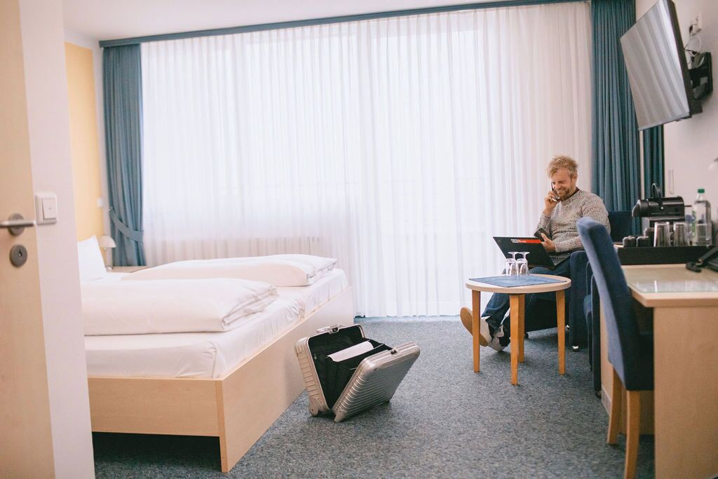 A hotel room with a bed, a desk, a chair, a small table, a suitcase, and a TV on the wall.