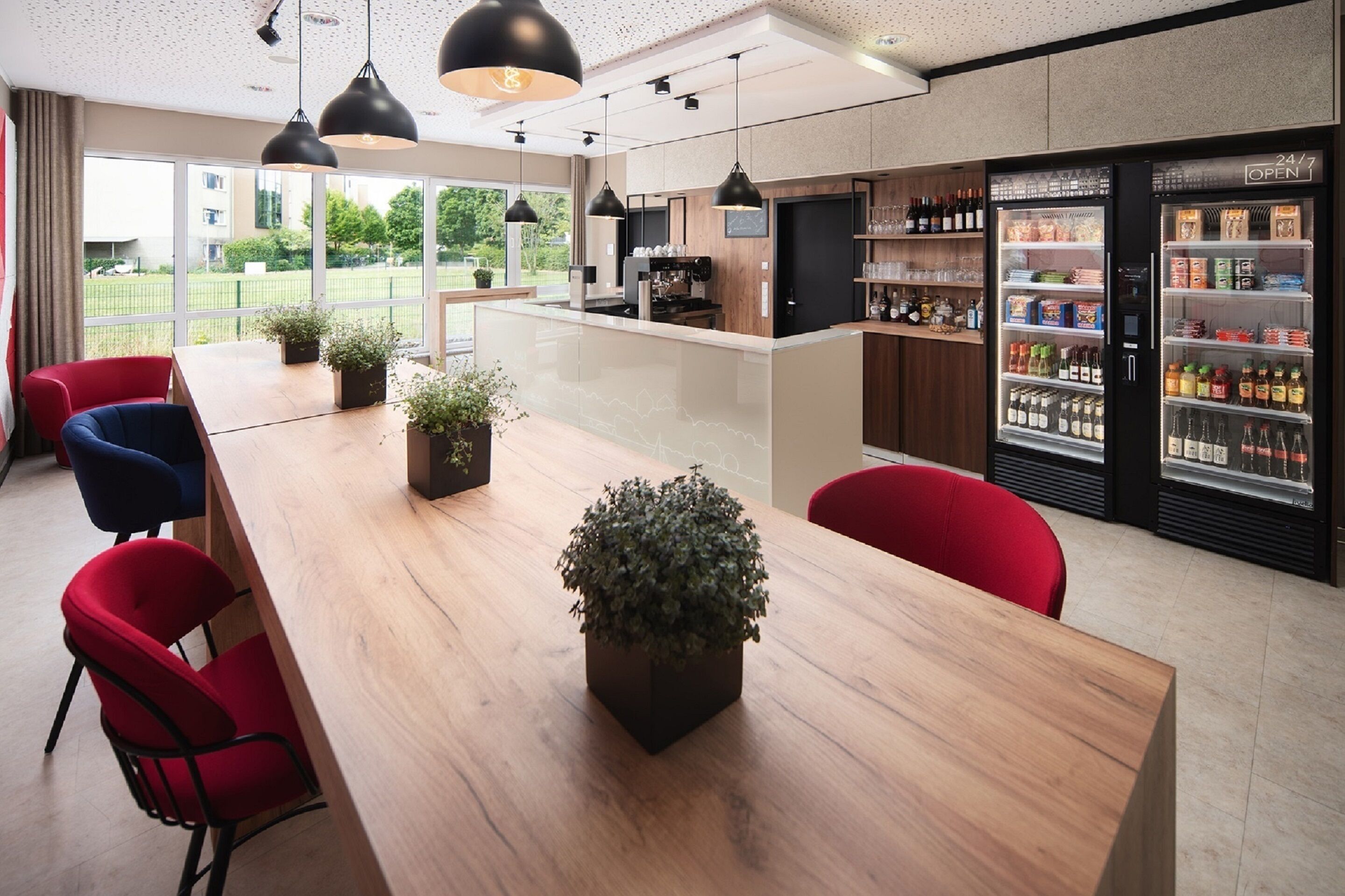 A modern hotel lobby with a long wooden reception desk, red and blue chairs, black pendant lighting, a coffee bar, and a refrigerated display case with beverages.