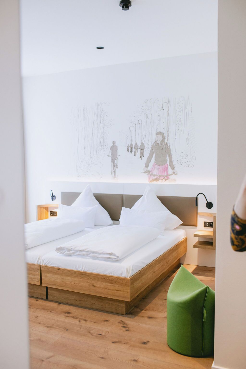 Ein modernes Schlafzimmer mit einem Holzbett, einem grünen Ottomanen und einem an der Wand montierten Projektor, der eine Szene zeigt.